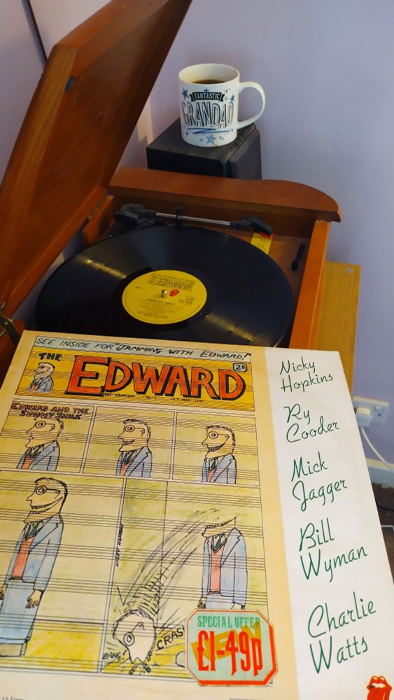 Record player with a 12" black vinyl record.
In the foreground is an album cover featuring a five panel cartoon strip titled "Edward and the Sunny Smile" in which a cartoon man's smile gradually widens until it separates the top of his head, which falls to the ground.
Behind the player is a mug of coffee on a speaker.