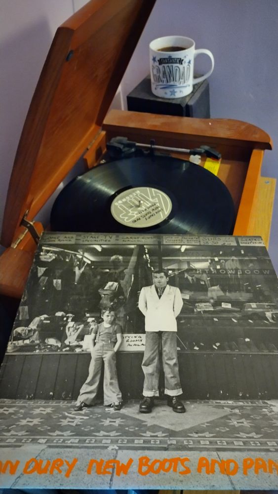 Record player with a 12" black vinyl record. In the foreground is an album cover showing Ian Dury and a young boy leaning against the front of a second hand shop.  Behind the player is a mug of coffee on a speaker.