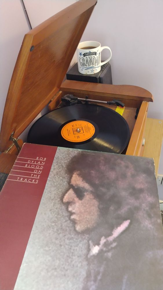 Record player with a 12" black vinyl record.
In the foreground is an album cover with a portrait painting of Bob Dylan in profile.
Behind the player is a mug of coffee on a speaker.