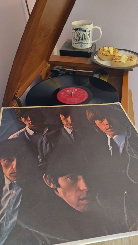 Record player with a 12" black vinyl record.
In the foreground is an album cover with a colour photo of the band, the right side of their faces in deep shadow, and all staring unsmiling directly at the camera.
Behind the player is a mug of coffee on a speaker.  On the corner of the player is a plate with two delicious buttered crumpets.