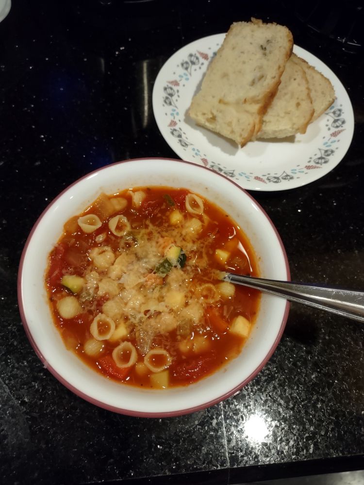 A white soup bowl filled with vegetable minestrone, topped with parmesan and a spoon, next to a small plate with rosemary focaccia.