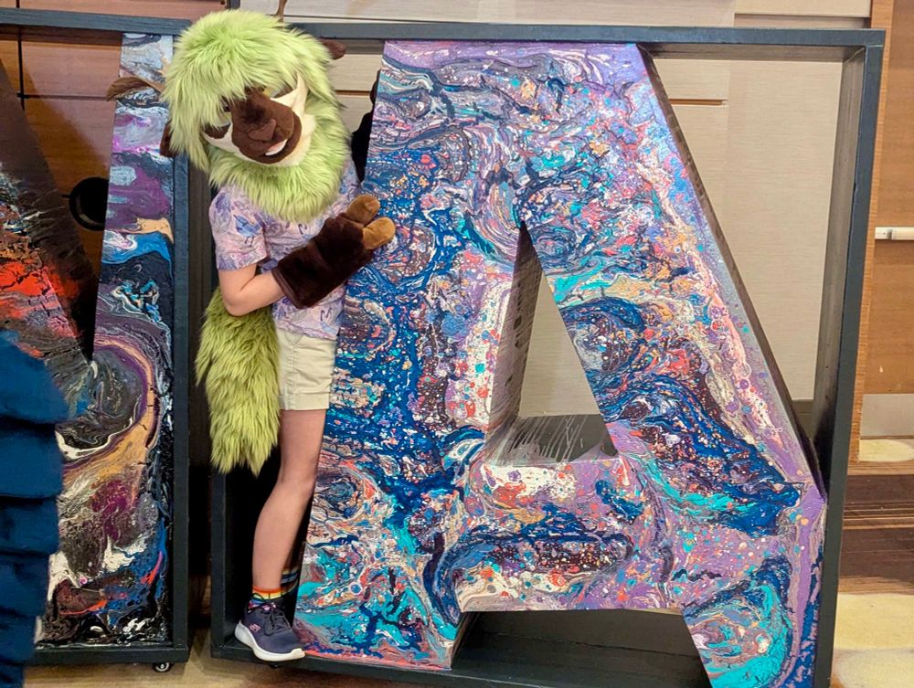 A green sheep fursona standing with a giant letter A statue with paint poured on it.