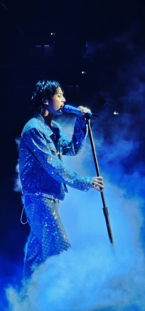 Jin standing and singing at the mic bathed in blue light and fog effects, wearing his sparkly denim on denim Gucci outfit
