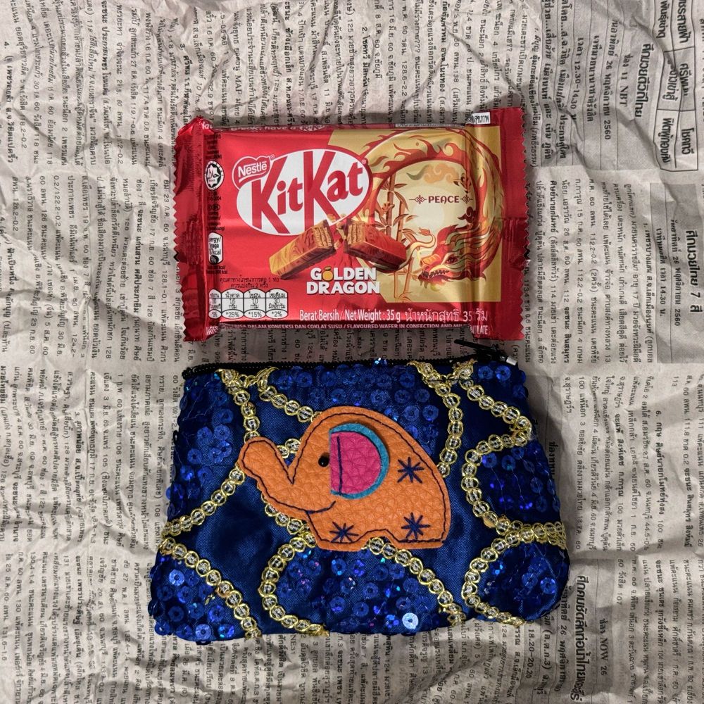 タイの新聞紙の上にキットカットとポーチが載っている写真。上はタイのキットカット、ゴールデン・ドラゴン味。下は象が縫ってある青色のスパンコールポーチ。