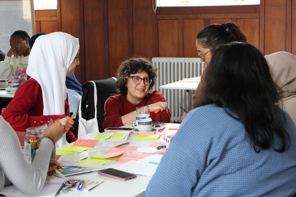 Sara Shaarawi crouches by a table covered in post it notes and cups of tea, talking to a group of young women. 