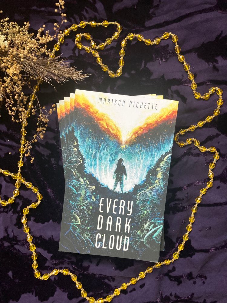 Photo of a stack of EVERY DARK CLOUD viewed from above, resting on a dark purple cut velvet background with a string of gold beads and dried flowers bordering the books. Cover art depicts a person standing silhouetted in blue-green light from bioluminescent mushrooms. Above, a minute figure falls through fiery clouds.