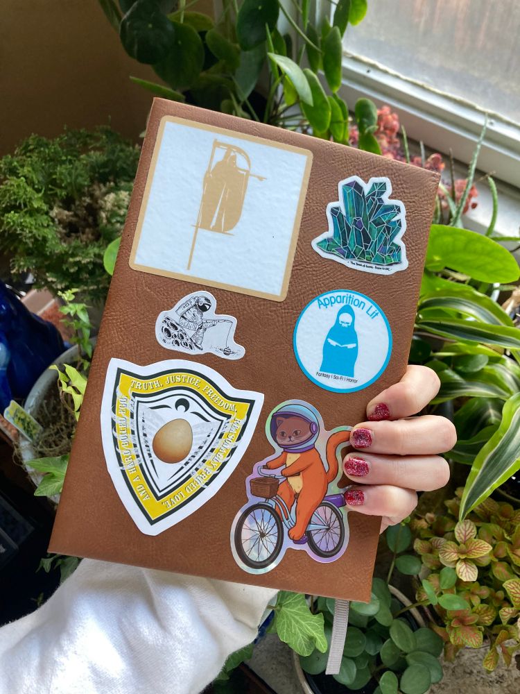 Photo of leatherette journal being held in left hand over potted plants. Stickers on back cover depict grim reaper, crystal cluster, astronaut fishing for planets, space cat riding bicycle, and shield emblem with “Truth, Justice, Reasonably Priced Love, and a Hard Boiled Egg” motto encircling an egg.