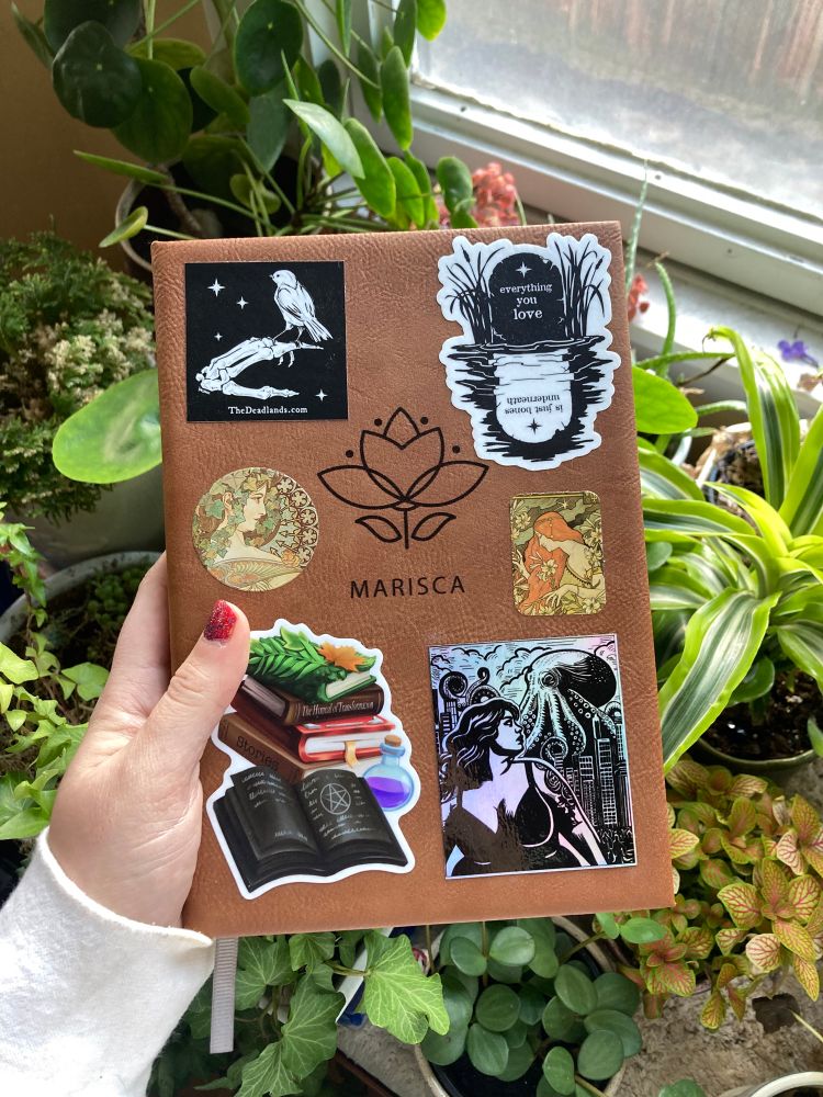 Photo of leatherette journal being held in left hand over potted plants. Stickers on cover include stack of spellbooks, art nouveau posters, skeleton hand balancing songbird, headstone, and octopus demolishing city with plus size woman in foreground.