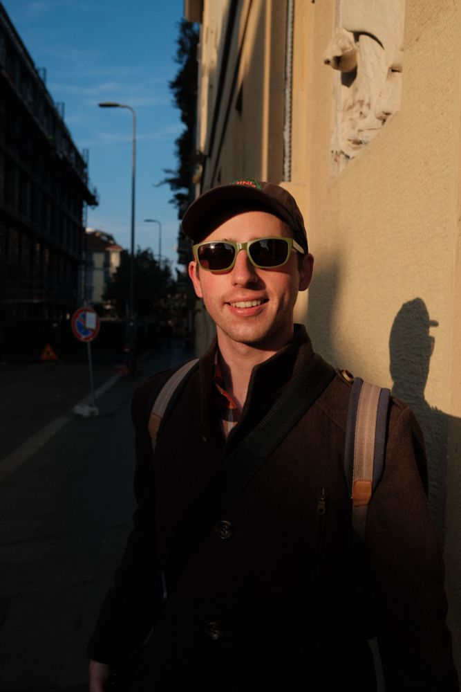 Photo of me on the street wearing a dark brown jacket, a backpack and with a camera by my side
