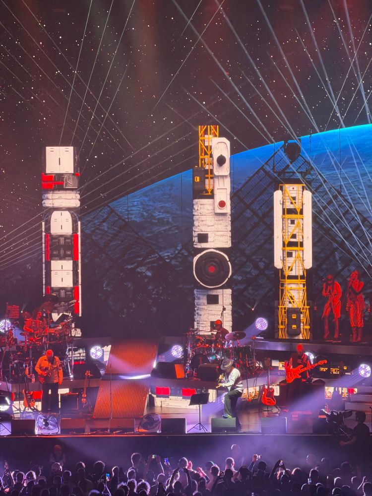 Stage above a crowd with musicians on it. Jay Kay is at the front in a white jacket and a hat. Behind the backdrop has rockets and the earth as if they were in space