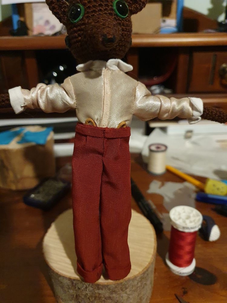 Front view of the doll. He is standing on a doll stand with his arms outsretched to the sides in a champagne coloured satin shirt featuring white cuffs and a white collar. The tiny shirt has a button stand down the front, and it's tucked into his rust coloured trousers. The trousers has suspender hoops on each side of the fly, and has a small pleat for fit going into the waistband. The hems are turned up on the outside in 1920's fashion.