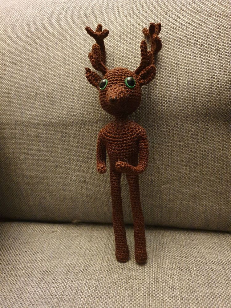 A crocheted doll standing upright. It is chocolate brown in colour with green safety eyes and big stag antlers, a muzzle and ears. It is masculine in stature, and the doll is simplistic in nature. 