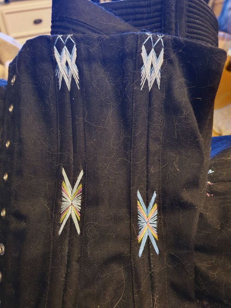 A close up of a black corset with flossing on top of four boning channels, which are done in three shades of blue and are criss crossing lines. On the middle of the boning channels there's also flossing, done in one shafe of blue, green and purple on the channels to the left, and on the right it has a darker shade of blue and pastel pink instead of purple. 