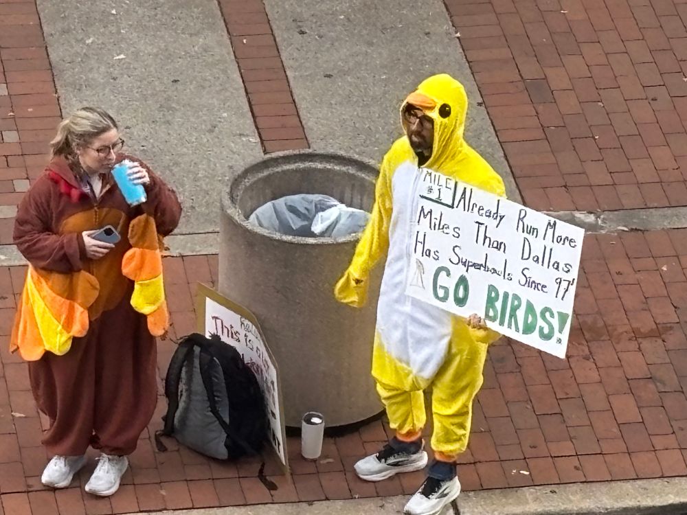 a man in a yellow bird costume holds a sign that reads “mile #1 already run more miles than dallas has superbowls since 97’ GO BIRDS”