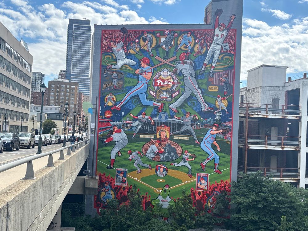the phillies mural on the side of a building includes numerous images of phillies icons, including: 1) Brad Lidge; 2) Charlie Manuel; 3) Mitch Williams; 4) Cole Hamels; 5) Grover Cleveland Alexander; 6) Larry Bowa; 7) Darren Daulton; 8) Dallas Green; 9) Tug McGraw; 10) Ed Delahanty (on the program cover for a game at Recreation Park); 11) Mike Schmidt; 12) Ryan Howard; 13) Chuck Klein (in front of the outfield wall at the Baker Bowl); 14) Tony Taylor; 15) Dick Allen; 16) Shibe Park (Connie Mack Stadium); 17) Jim Bunning; 18) Veterans Stadium; 19) Roy Halladay; 20) Citizens Bank Park; 21) Robin Roberts; 22) Harry Kalas; 23) Richie Ashburn; 24) Steve Carlton; 25) Jimmy Rollins; 26) Chase Utley; 27) Johnny Callison; 28) Curt Simmons; 29) Del Ennis; 30) Greg Luzinski; 31) Carlos Ruiz; 32) The Phillie Phanatic