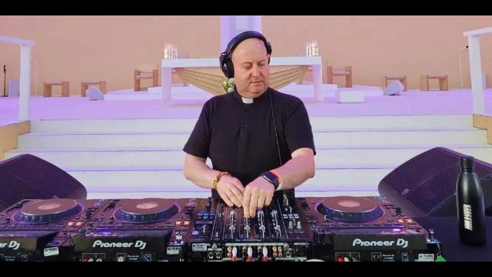 dj padre guilherme in a black short sleeve shirt with a roman collar standing at turntables 