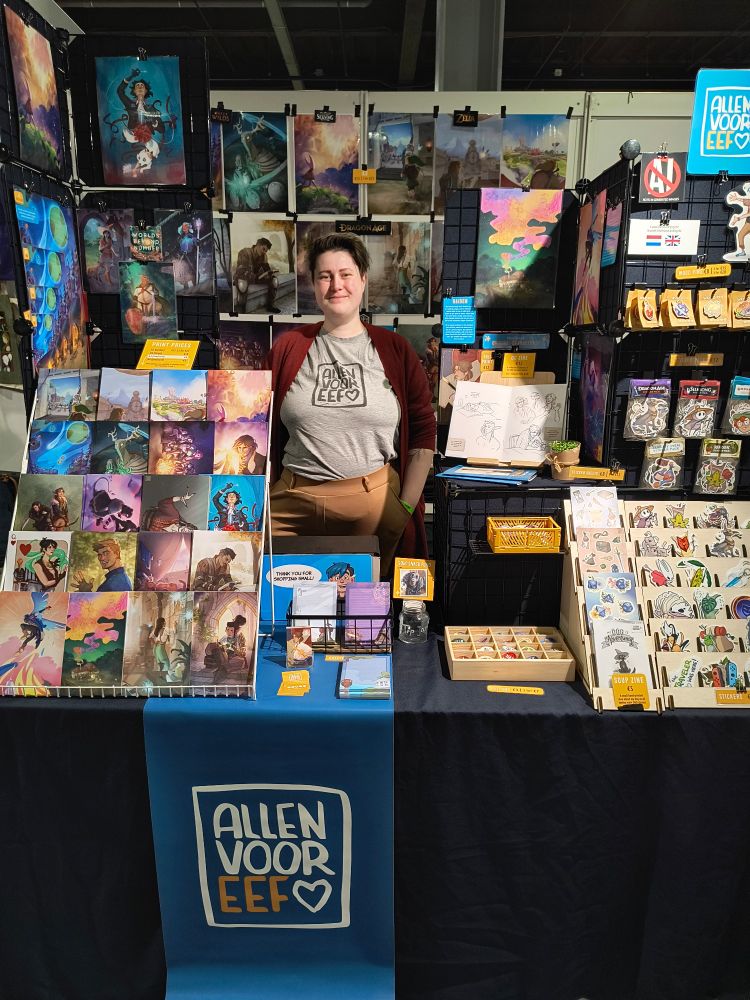 A picture of me behind my Dutch Comic Con table, filled with art prints and merch.