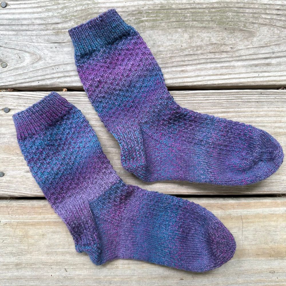 Photo: a pair of purple and blue handknit socks lying on the weathered boards of a deck. 