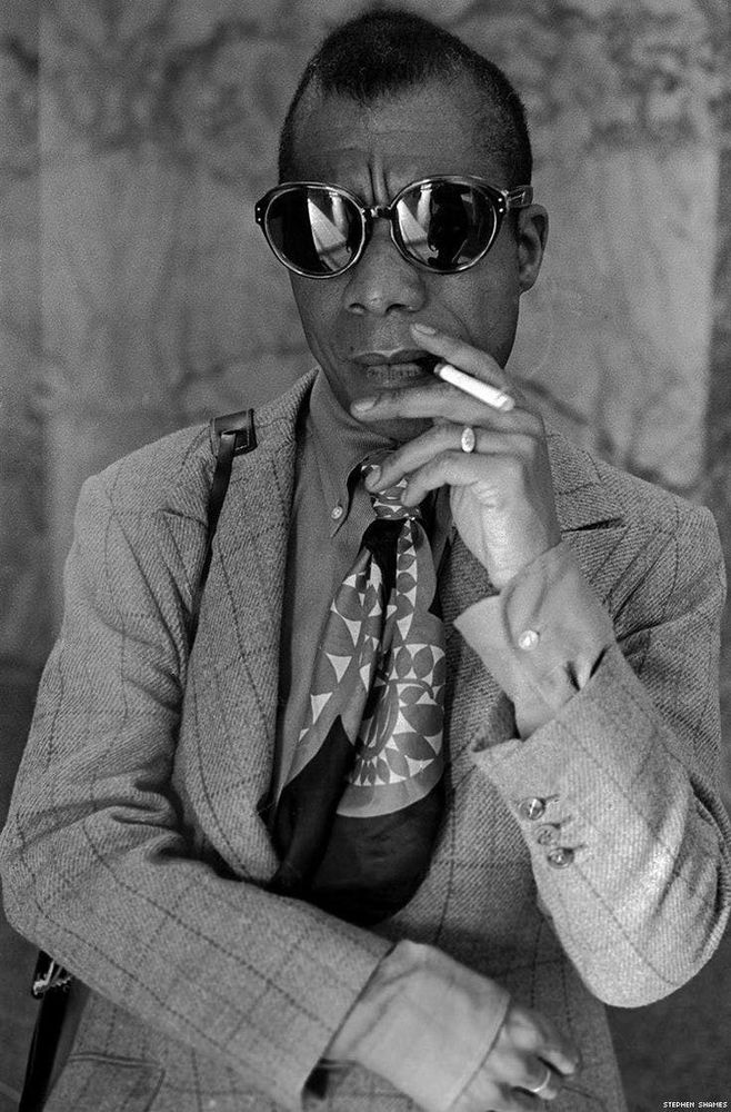 Black and white photo of James Baldwin. He is wearing a tweed jacket and dress shirt underneath, a silk scarf cravat, slightly oval sun glasses, and is smoking a cigarette. There is a black bag strap over his right shoulder and he is wearing a ring on his left ring finger.
