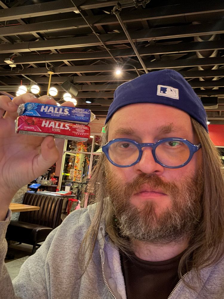 poster in backwards baseball cap and light blue glasses, holding up red & blue HALLS brand cough drops