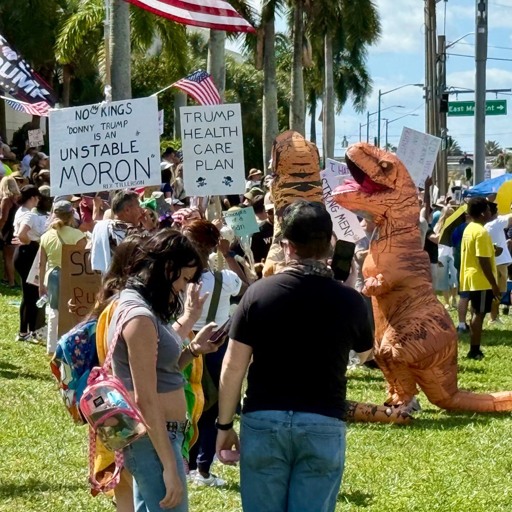 Crowd gathered near intersection includes two participants wearing inflatable t-rex costumes. Others hold posters reading, “No kings. Donny Trump is an unstable moron” and “Trump healthcare plan” with two skull and crossbones symbols. 