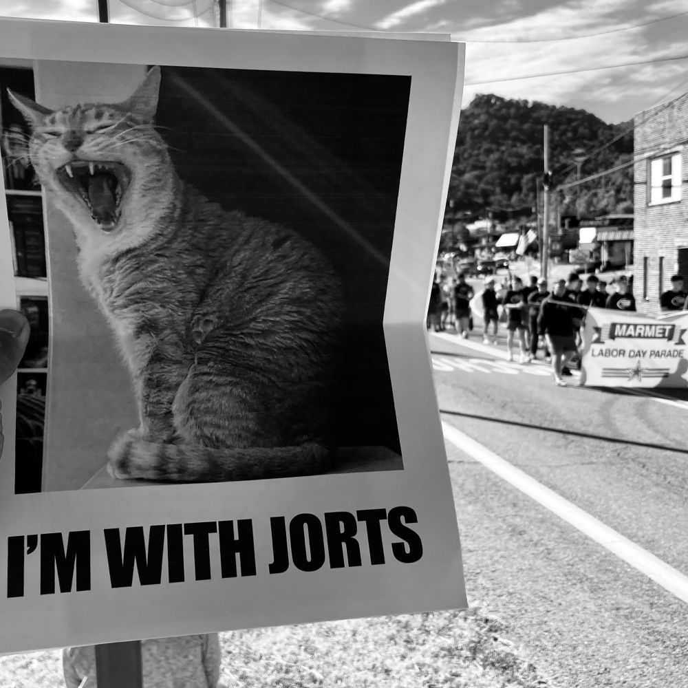 Flyer of Jorts the Cat with Marmet Labor Day Parade participants carrying a banner in the background. 