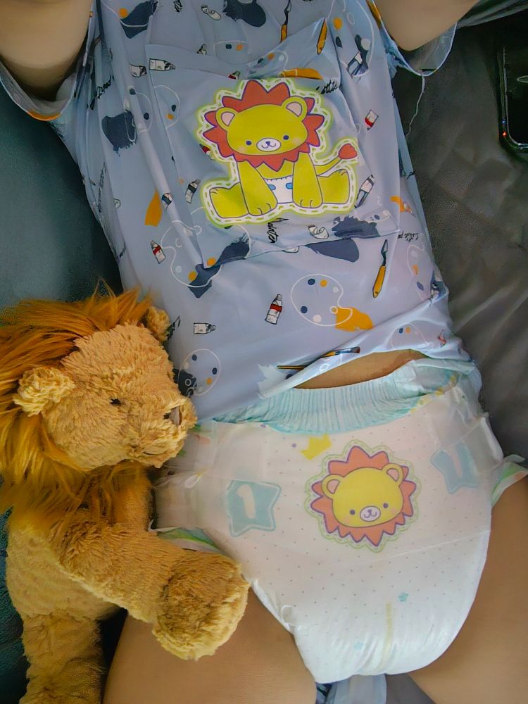 That lioxy kiddo lying in bed wearing a hoodie onesie with the ABU Little Kings lion printed on and an ABU Little Kings diaper, with their lion plushie Maximilian next to them