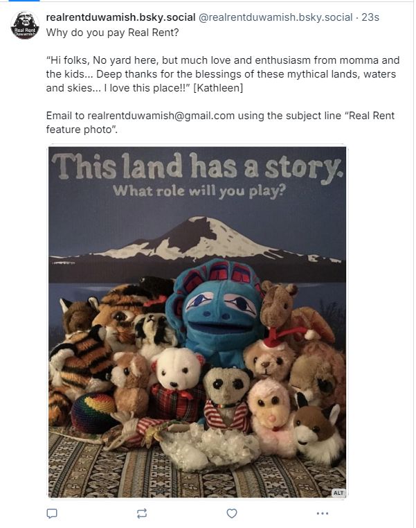  Blue Duwamish yard sign with Mount Tahoma (known by settlers as Mount Rainier) portrayed in black and white and the words “This land has a story. What role will you play?. In front of the sign is a group of stuffed animals and a large quartz crystal.