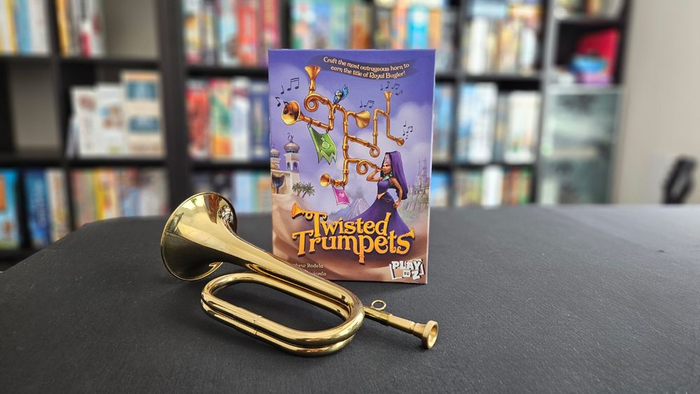 Shot of the Twisted Trumpets board game box with a bugle sitting next to it and a boardgame shelf in soft focus in the background.