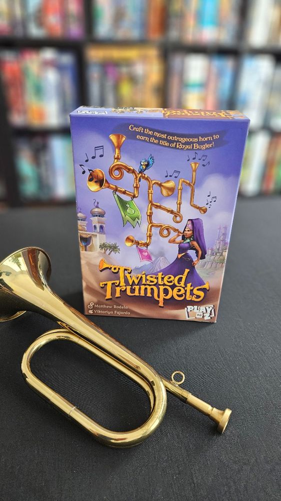 Shot of the Twisted Trumpets board game box with a bugle sitting next to it and a boardgame shelf in soft focus in the background.