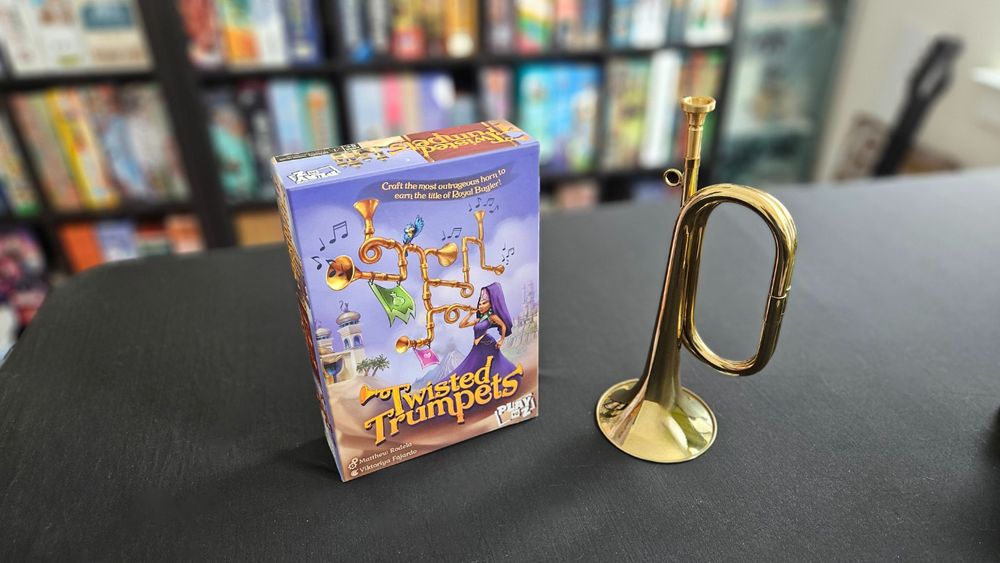 Shot of the Twisted Trumpets board game box with a bugle sitting next to it and a boardgame shelf in soft focus in the background.
