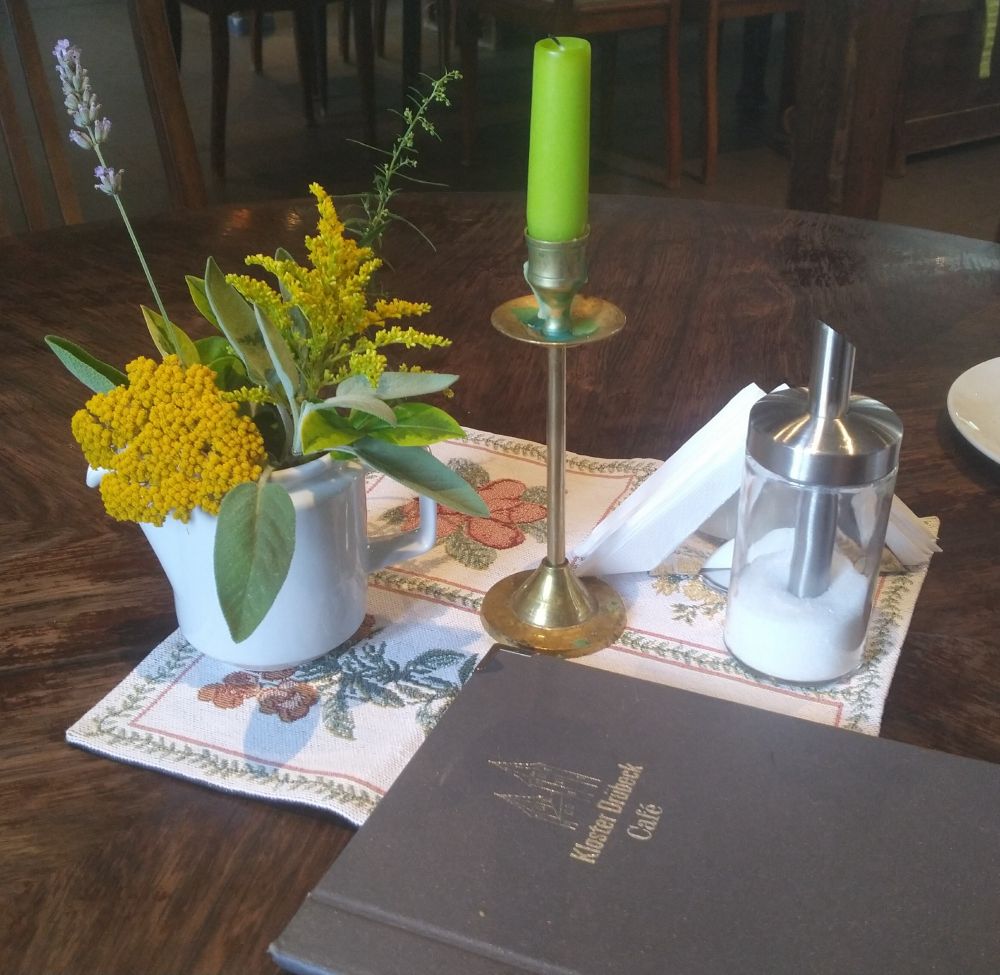 decoratie arrangementje van verschillende gele (wilde) bloemen, lavendel en groen/gele takjes met groene kaars op koperen kandelaar, glazen suikerpotje met rvs giettuitje, rvs servettenstandaard samen op een bont kleedje op ronde donkerbruine eiken tafel in kloostercafé Drübeck, Ilsenburg, Harz (ST)