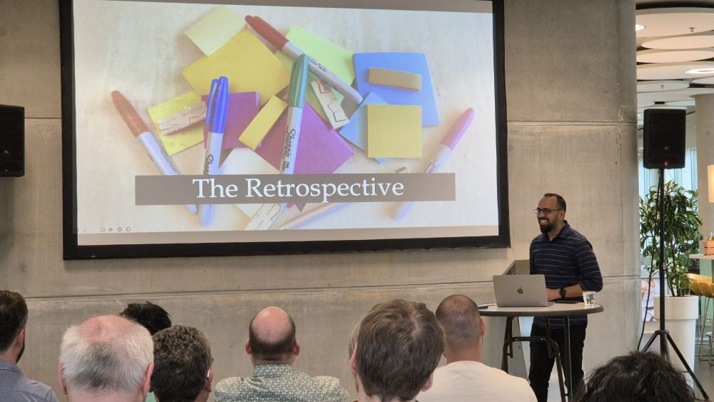 Rijo Sam smiling and telling stories about "Tulips to Turmeric" in front of a big screen stating 'The retrospective' with an image of sticky notes and blue pens. In front of the JUGNoord attendees.