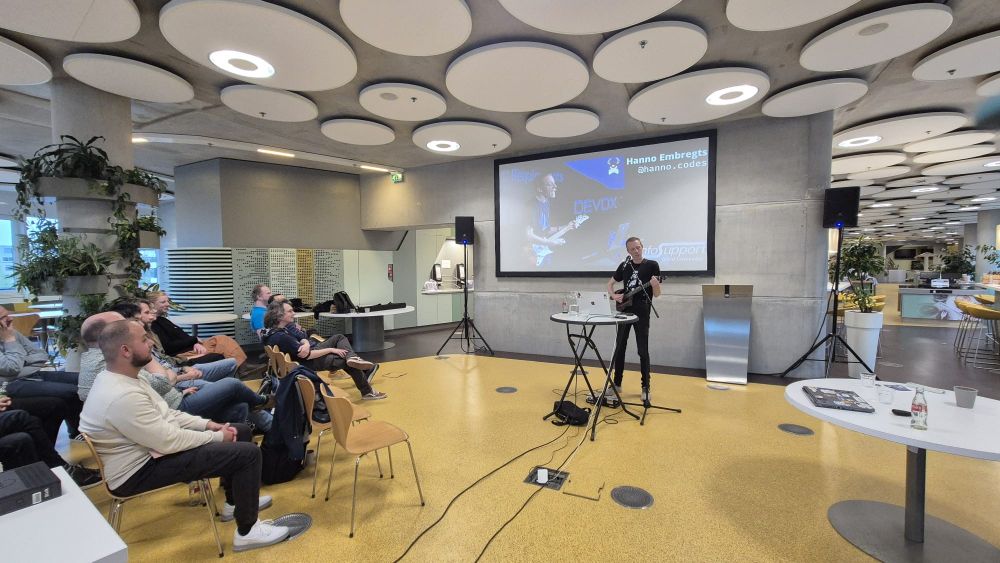 Hanno Embregts playing guitar. In front of him is a music standard for the microphone and a high table with an apple laptop on it. Behind him is a big screen with a cool photo that was taking during his key note at Devoxx Belgium a couple years ago. On the left you see some attendees from the JUGNoord event in the DUO building.
