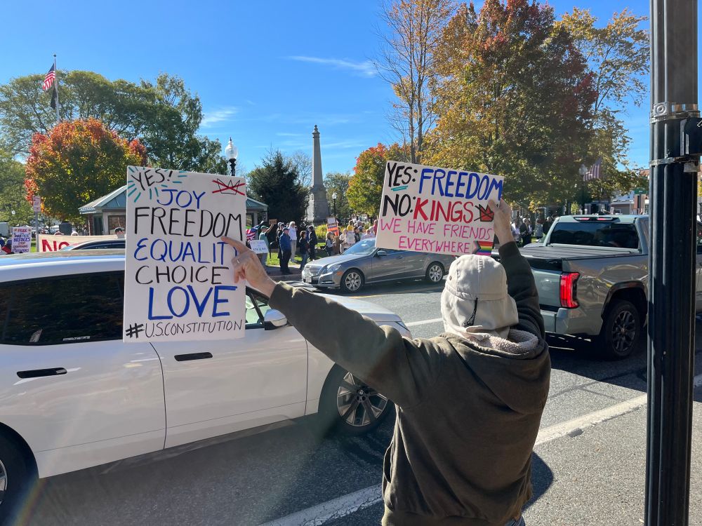 A man in a sun hat and hoodie holds two signs as cars pass. Across the street is a crowd with other signs. His signs read “Yes: Joy, freedom, equality, choice, love, #USConstitution” and yes: freedom No:kings, We have friends everywhere with a badly drawn but heartfelt pride flag. 