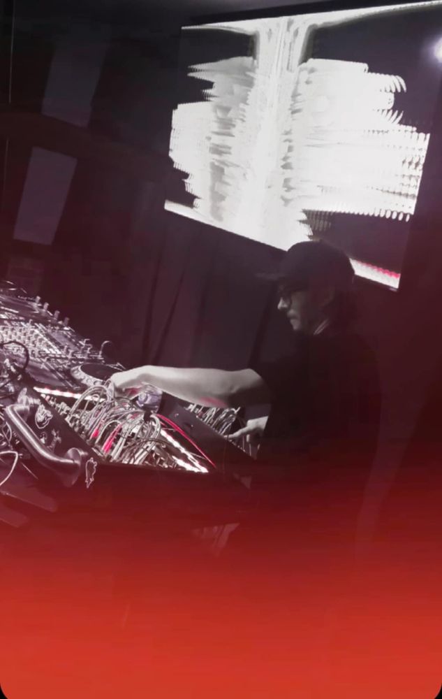 me, operating a modular synth system, next to a DJ mixer and decks, large TV with visuals behind me