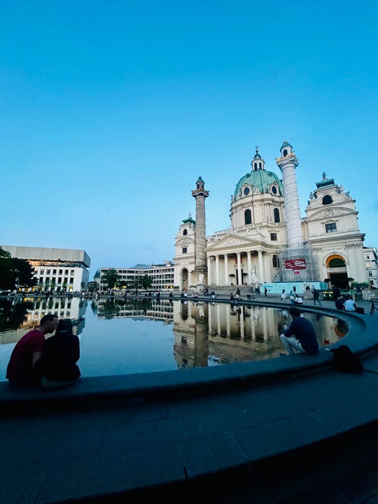 Abendstimmung am Karlsplatz in Wien. Im Vordergrund sitzen Menschen am Rand des Wasserbeckens, das die barocke Karlskirche mit ihren markanten Säulen und der grünen Kuppel perfekt spiegelt. Der Himmel ist klar und blau, das Licht sanft und kühl. Rund um das Becken verweilen Menschen, manche allein, manche in Gespräche vertieft. 