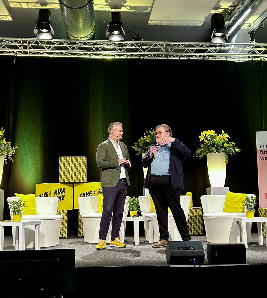Patrick Rammerstorfer im Gespräch mit Wolf Lotter auf der Bühne des „Festival of Curiosity“ in der Lösehalle der Tabakfabrik Linz. Beide stehen vor einem Hintergrund aus grünen und gelben Farbtönen mit Blumentöpfen, Scheinwerfern und Dekorationen. Wolf Lotter spricht in das Mikrofon, Patrick Rammerstorfer hört zu.