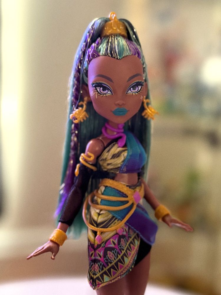 Monster High doll Nefera de Nile with long teal and purple streaked hair styled in a high ponytail, golden crown-like hairpiece, and bold turquoise lipstick. She wears a gold and teal asymmetrical outfit with geometric patterns, gold jewelry, and a purple sash, posing with one arm slightly raised.
