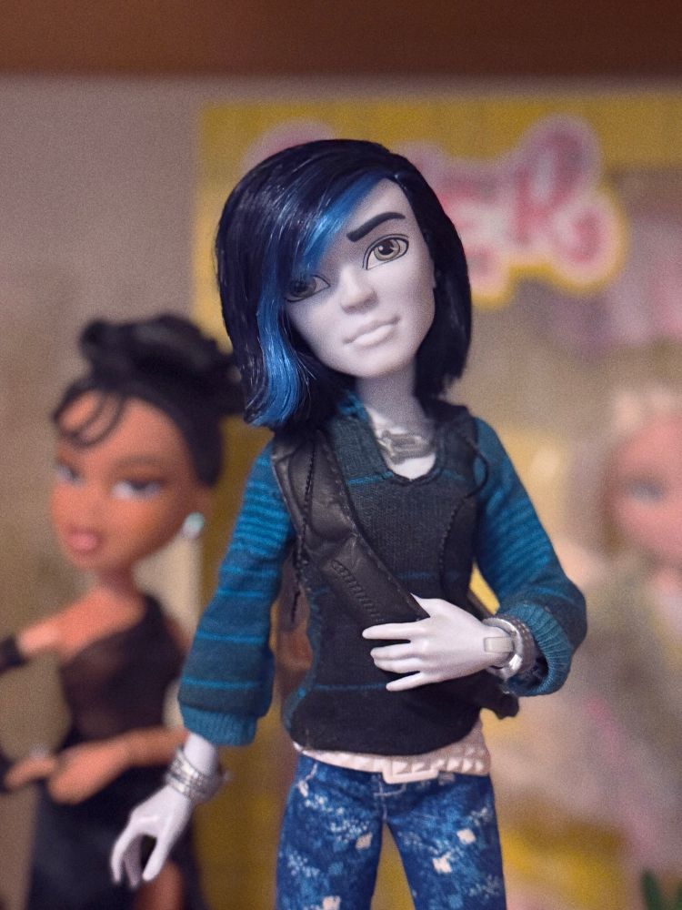 A photo of a monster, high doll composed in the centre of the shot on the shelf in front of two bra stalls blurred on a shelf probably belonging to a collector the monster high doll has white skin blue hair with a light blue streak. He’s wearing a striped blue sweater and printed blue shorts and a white belt with silver jewelry and a black satchel backpack.