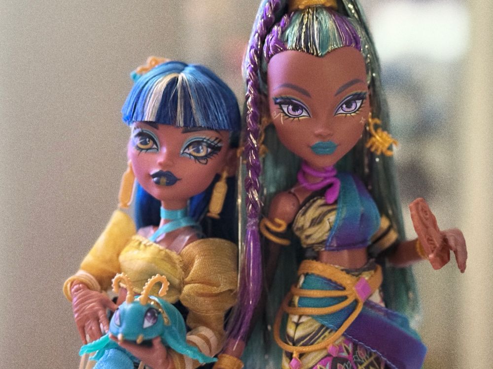 Monster High sisters Cleo de Nile and Nefera de Nile posed together. Cleo, on the left, has straight blue hair with blunt bangs, wears a gold and black outfit with patterned details, and holds a small turquoise creature with gold accents. Nefera, on the right, has teal and purple streaked hair in a high ponytail, turquoise lipstick, and a gold-and-teal patterned outfit with a purple sash, holding a small brown accessory.