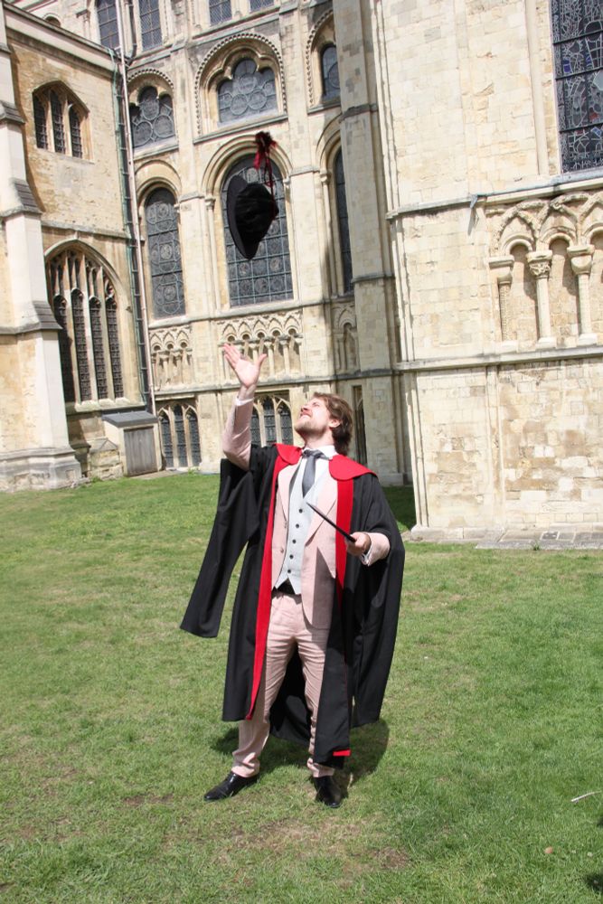 A man outside Canterbury Cathedral in a doctoral gown holding a degree certificate and throwing his hat into the air. 