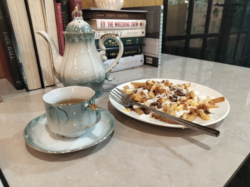 Matching Tea pot and tea cup,  a white plate of French fries with a  fork, and a stack of book on the background.