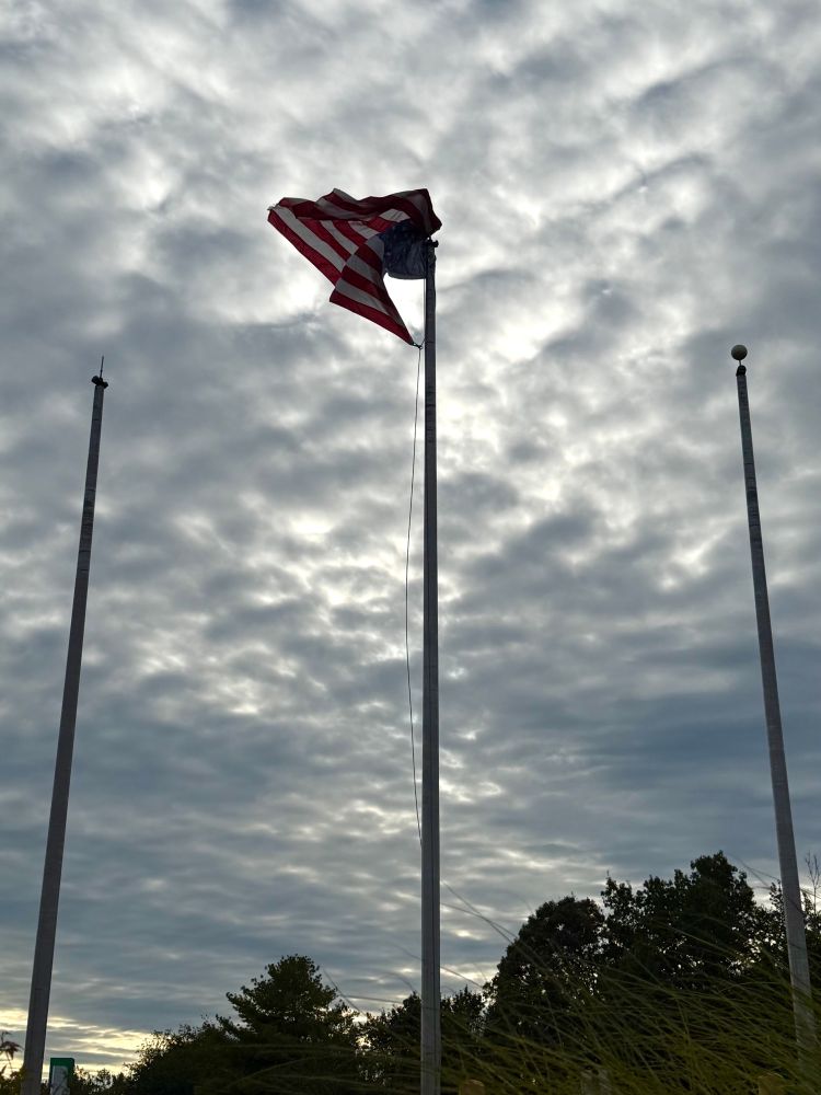 a tangled US flag on a flag pole in front of a cloudy sky. something has come loose and it's flipped over the top of the pole, so now it's wadded up and looking the worse for wear