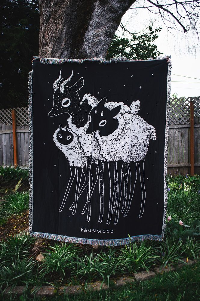 A photo of a blanket tapestry! It is 100% woven with a blend of cotton and recycled cotton, made in the US.  The design is black and white with three woolly deer creatures.  The middle creature is covered in stars, and there are some stars in the background too.  The text on the bottom says “Faunwood”.  