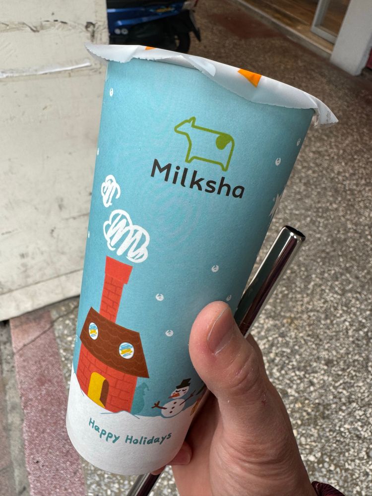 Vertical photo of a hand holding a Milksha takeout cup of pearl milk tea or boba tea. The cup art shows a winter cartoon scene with a snowman and smoky chimney with tagline Happy Holidays at bottom, completing the irony of trendy marketing in Taiwan as a subtropical country with neither deep snowy winters nor majority Christian/Germanic traditions.