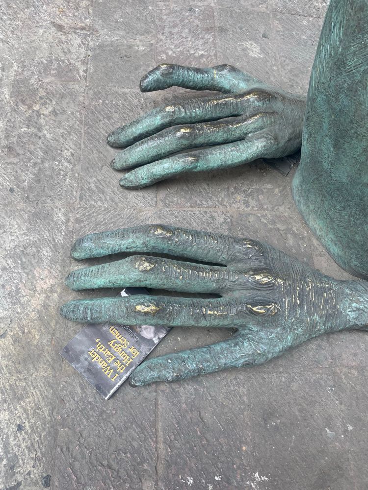 A copy of my book, I Wander the Earth, Hungry for Semen, peeking out from beneath a sculpture/bench. The sculpture looks like a bird but has four fingered hands. 