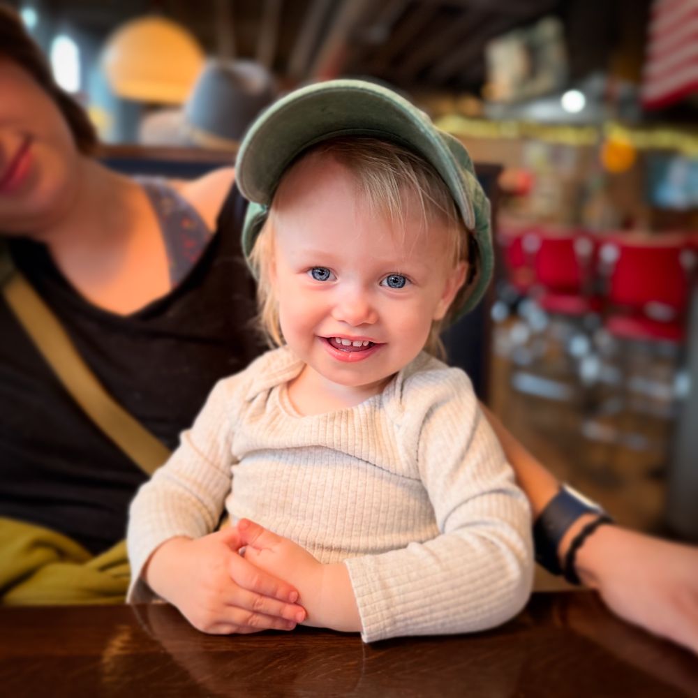 Adorable toddler girl smiling at camera while wearing dada’s hat and sitting on mama’s lap in booth at restaurant