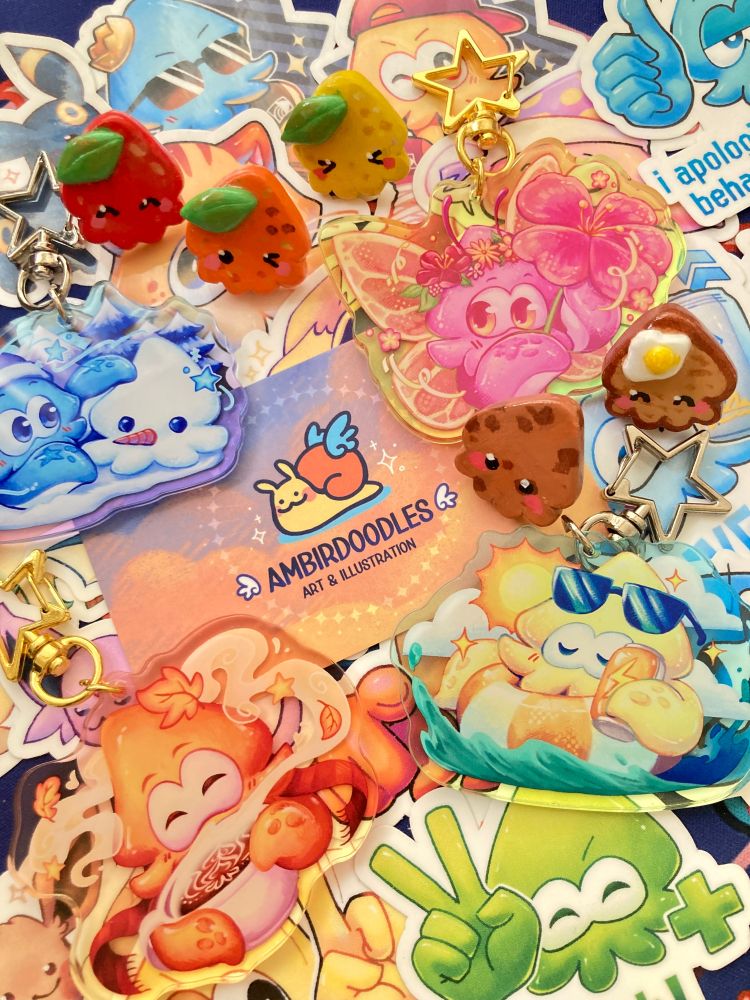a photo of a collection of ambird's stickers, charms, and pins scattered on a table. in the middle is a business card with their logo and name on it.