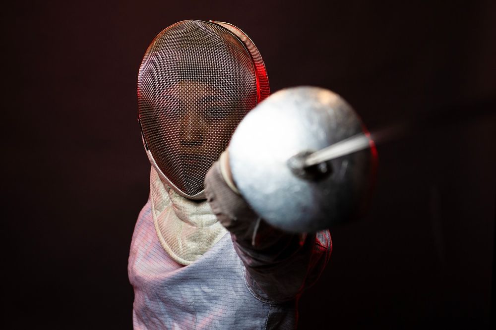 A student on the fencing team, who is dressed in fencing gear, points their sword at the camera.